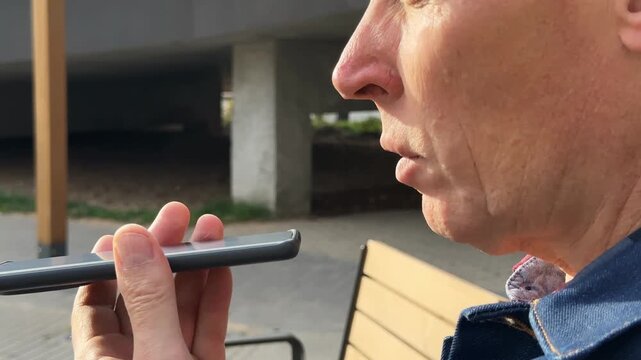 Mature man recording audio voice message on smartphone device outdoors. Senior male person holds phone near face to dictate vocal note in messenger app. Voice recognition technology allows