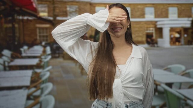 Young woman in white blouse covers eyes with hand while smiling beside outdoor cafe tables and chairs on a street near brick building; playful.