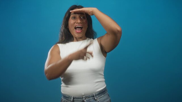 Young hispanic woman shielding eyes with hand and pointing while wearing a white tanktop and jeans against a blue studio backdrop; curiosity discovery.
