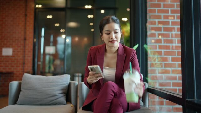 Young businesswoman using her smartphone and drinking matcha green tea in a modern office lobby or cafe during a work break