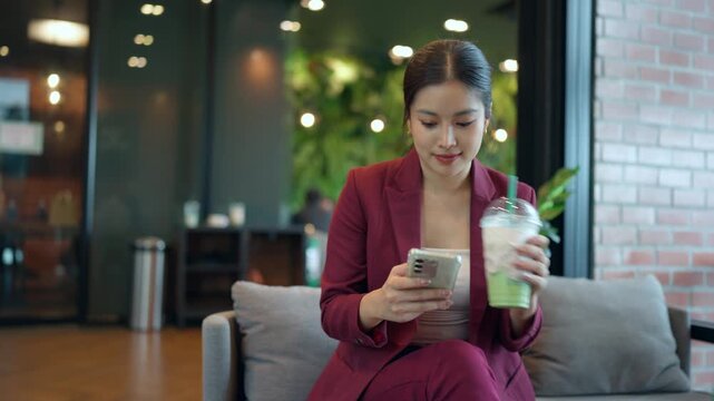 Asian businesswoman using a smartphone and drinking iced green tea latte while sitting in a modern office lobby or coffee shop
