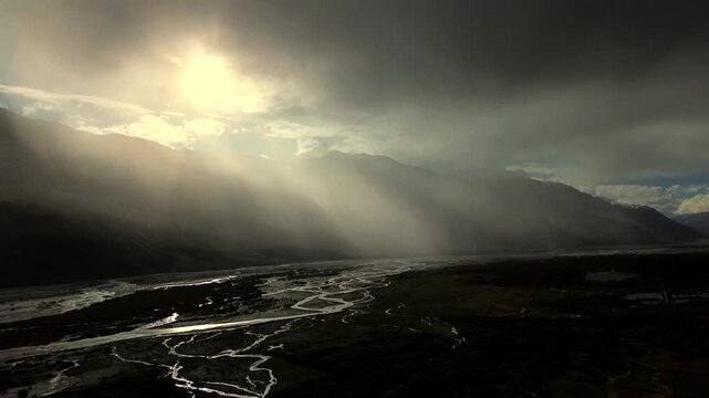 Wide high angle aerial pan right shot capturing dramatic sun rays breaking through storm clouds over the reflective braided river in Nubra Valley, Ladakh, India.
