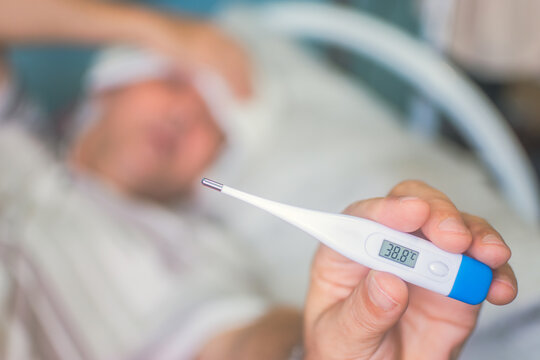 A hand holds a digital thermometer showing a high temperature. In the blurred background, a person lies in a bed, appearing ill.
