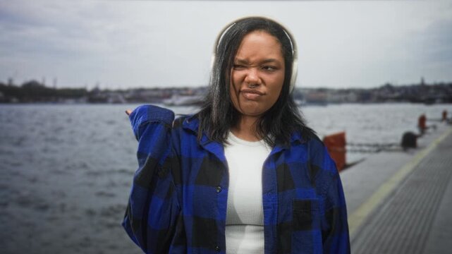 Young woman with headphones shrugs and grimaces, hand to ear on a waterfront street pier; annoyed skeptical.