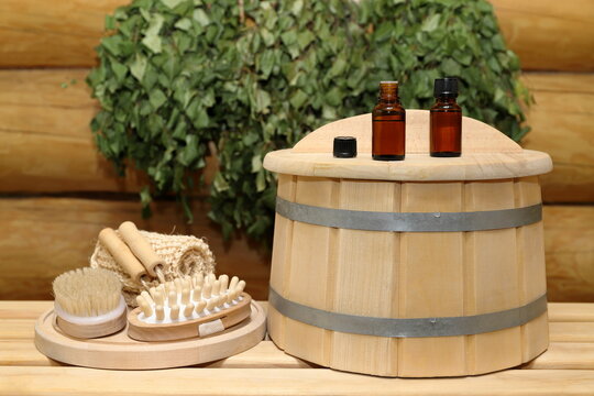 On a plank bench, on top of a wooden barrel, next to set of bath supplies on wooden round tray, there are two bottles of healing essential oil, against the background of log wall with a birch brooms. 