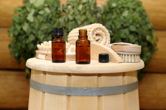 On top of a wooden barrel, next to a set of bath supplies, there are two bottles of healing essential oil, against the background of a log wall with a birch brooms.