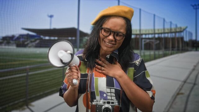 Young woman filmmaker wearing yellow beret and glasses holds megaphone, hand on chest and eyes closed, vintage camera at neck on street beside stadium stands; reflective resolve.