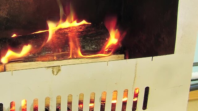 The footage shows burning wood in dark metal fire chamber. The frame also shows orange flames burn around dark wooden logs inside dark metal chamber within dark metal fire chamber.