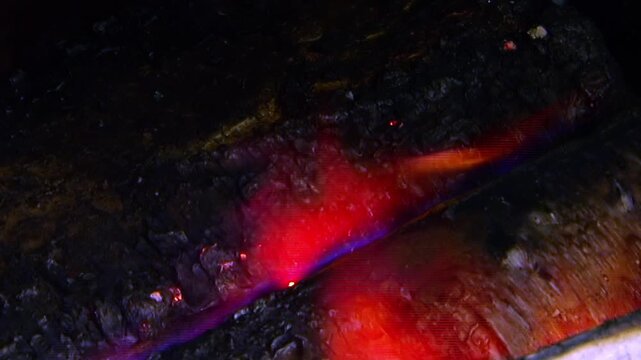 The footage shows burning wood in vented metal chamber. The shot also includes orange flames burn around dark wooden logs beside metal ventilation slots across the visible setting.