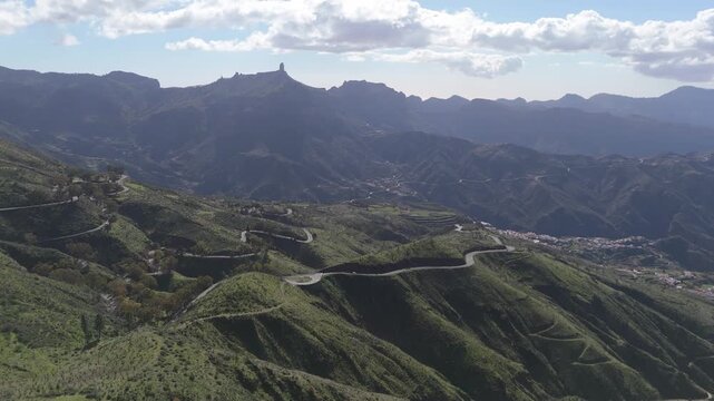 Aerial drone push-in shot revealing winding mountain roads, lush green hills, and volcanic peaks on Gran Canaria, Canary Islands, Spain. A remote and scenic island landscape.