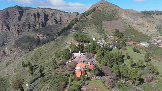 An aerial drone shot of the picturesque Cruz de Tejeda hamlet and its surrounding mountainous landscape on Gran Canaria, Spain. The drone pushes in, revealing the lush hills, roads, and buildings.
