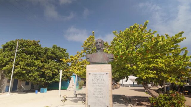 Dolly out revealing bronze bust monument of Simon Bolivar, the Liberator, mounted on stone pedestal in town square of Gran Roque island. Lush green tropical trees and sandy ground