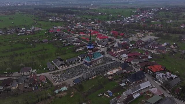 Drone view over the colorful church and cemetery in Săp&acirc;nța, Romania, surrounded by village houses, green fields and the rural landscape of Maramureș