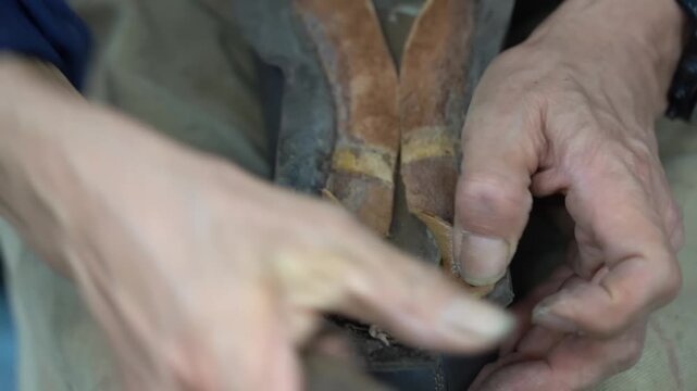 Craftsman trimming the excess welt of a dark leather boot with a sharp knife and pliers across his knee