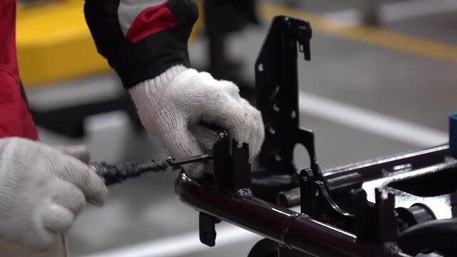 Assembly worker in a red uniform and white gloves tightening screws on a dark red brush cutter frame with a pneumatic screwdriver