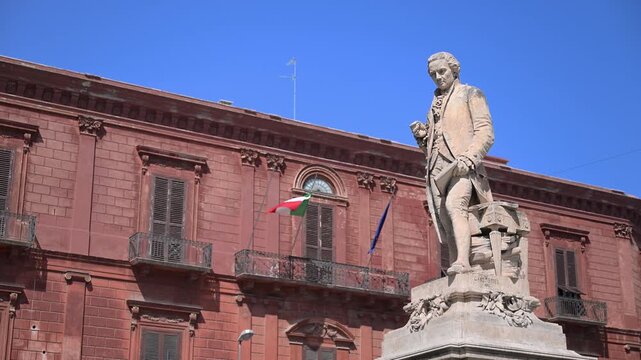 Marble statue of Niccolo Piccinni stands before a red historic building with Italian and EU flags against a clear blue sky.