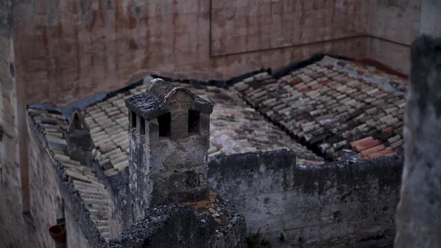 Old stone chimney and uneven terracotta roof tiles in Matera come into focus in soft evening light.