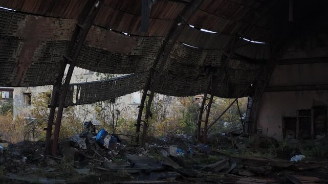 Slow right pan across the interior of a dilapidated industrial building with a rusted, sagging metal roof, revealing a forest and clear blue sky through a large open doorway.