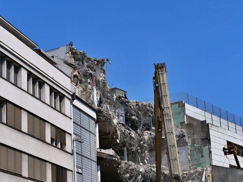 Heavy Demolition Excavator Arm Dismantling Concrete Building Wall Next to Office Building on Urban Construction Site