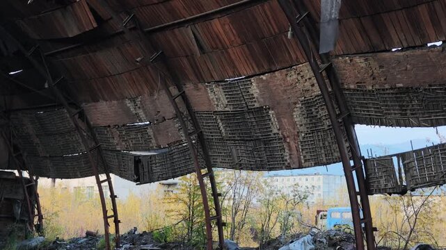 Slow pan beneath a dilapidated, rusted metal roof with sagging panels reveals autumn trees, distant buildings, and a blue vehicle on the ground under a clear blue sky.