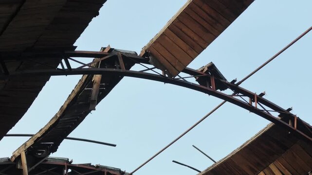 A deteriorating wooden roof structure with exposed beams and missing planks reveals a clear blue sky as the camera slowly pans downward to highlight warped slats and rusted metal supports.