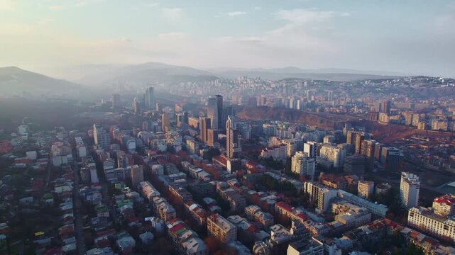 flight over Tbilisi Mixed urban architecture recedes during a smooth drone retreat from red roofed streets and long apartment rows modern towers dense hillside housing and mountains softened by haze