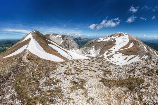 View of the glacial Lago di Pilato nestled between the snow-capped peaks of the Sibillini Mountains under a clear blue sky in Montemonaco, Marche, Italy.