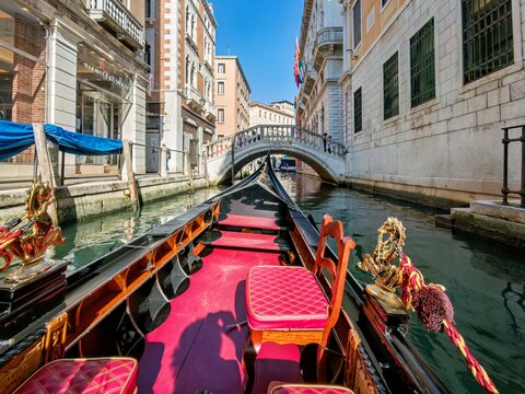View of a traditional gondola with red velvet seating navigating a narrow canal surrounded by historic stone buildings and a small arched bridge in Venice, Veneto, Italy.