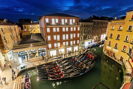View of gondolas moored in a canal at night with illuminated historic buildings and reflections in the water in Venice, Veneto, Italy.