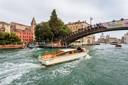 View of a wooden motorboat cruising on the Grand Canal with the Accademia Bridge and historic buildings in the background in Venice, Veneto, Italy.