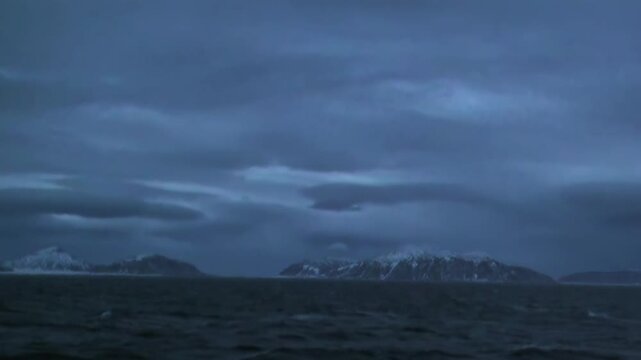 The footage shows dark coastline and water in open sea. The composition includes dark blue gray water with low mountainous land beneath cloud cover across the visible setting.
