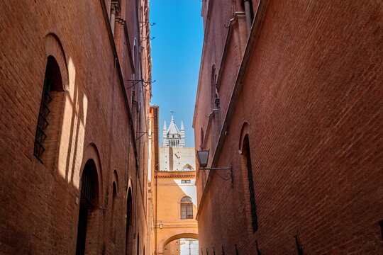 View of the Siena Cathedral tower visible through a narrow medieval brick alleyway in the historic center of Siena, Tuscany, Italy.