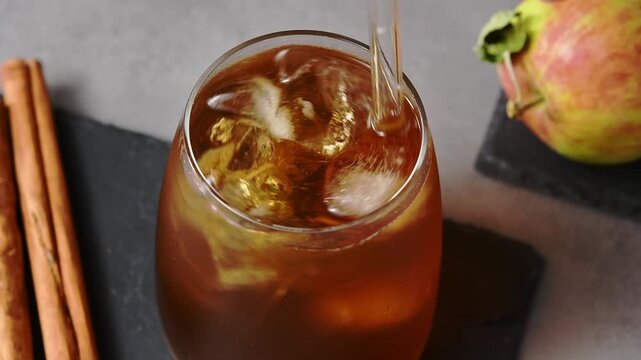 Hand stirs apple cider cold brew tonic with glass straw in close view beside apples and cinnamon.