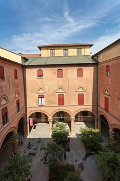 View of the historic courtyard of Palazzo d'Accursio featuring red brick architecture, arched porticos, and small trees in Bologna, Emilia-Romagna, Italy.