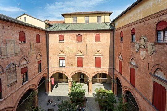 View of the historic courtyard of Palazzo d'Accursio featuring red brick architecture, arched porticos, and small trees in Bologna, Emilia-Romagna, Italy.