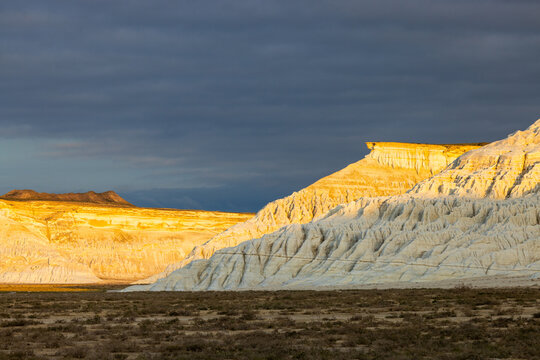 View of the white chalk cliffs of Tuzbair illuminated by golden sunlight against a dark stormy sky in the desert landscape of Mangystau District, Mangystau Region, Kazakhstan.