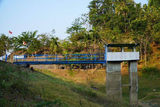 Blue bridge spans green hillside. Concrete pillars support its structure. Red flag waves gently in breeze. Trees tower behind the bridge. Sunlight filters through dense canopy