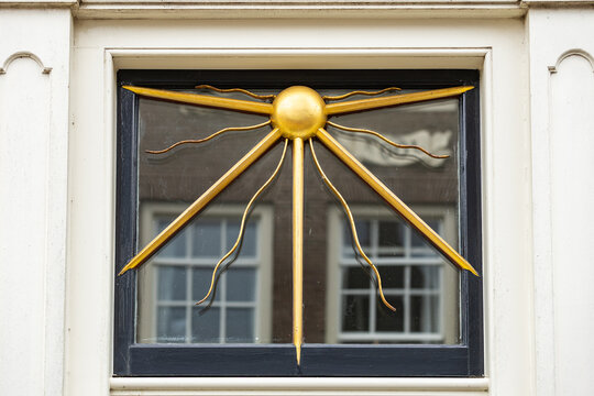 View of a gold sunburst ornament mounted on a window frame with white trim and reflections of nearby buildings in Monnickendam, North Holland, Netherlands.