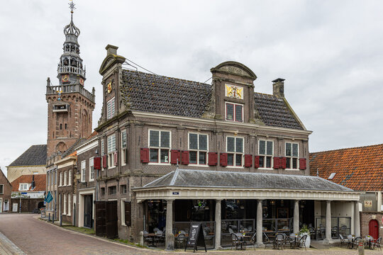 View of the historic Waag building with ornate facade and red shutters, featuring a church tower in the background under a cloudy sky in Monnickendam, North Holland, Netherlands.