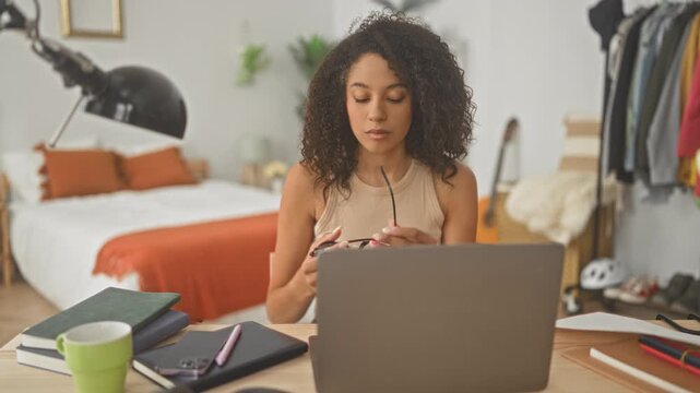 Woman typing on laptop and adjusting glasses, hands on keyboard in a bedroom workspace inside a residential building; focused study calm.
