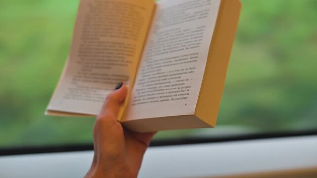 A person holds a book open while seated on a train, with pages visible. The background shows greenery through the train window, indicating travel through a natural setting