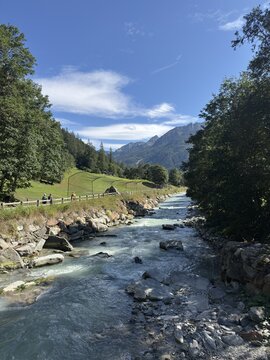 View of the Lys river flowing through the valley with people walking on a path, lush trees, and mountains under a clear blue sky in Gressoney-Saint-Jean, Aosta Valley, Italy.