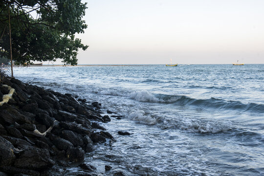 View of the rocky coastline of Saint Martins Island with waves hitting dark stones, foliage, and small fishing boats on the sea in Cox's Bazar District, Chittagong Division, Bangladesh.