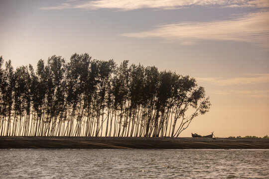 View of Shah Porir Dwip with a line of trees along the sandy shore and a small boat resting on the beach under a soft golden sky in Dakhinpara, Chittagong Division, Bangladesh.