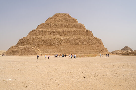 View of the Step Pyramid of Djoser in the desert with tourists walking in the foreground under a clear sky in Badrshein, Giza Governorate, Egypt.