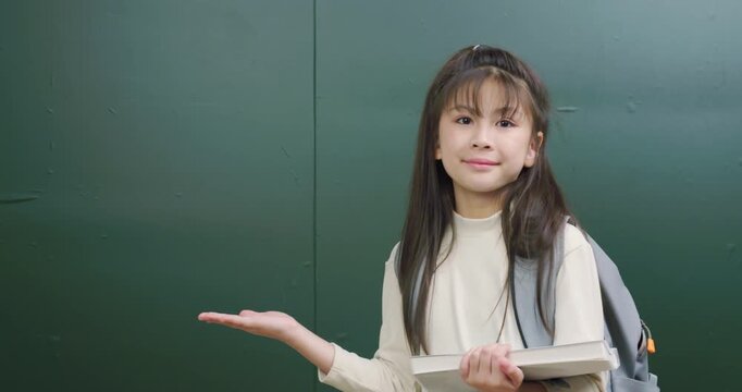 Cute Asian student girl holding book and presenting empty space against green background.