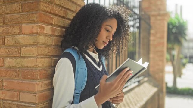 Woman reading a hardcover book, hands holding pages and turning a bookmark, wearing school uniform and backpack against a sunlit brick building on a city street; contemplation study.