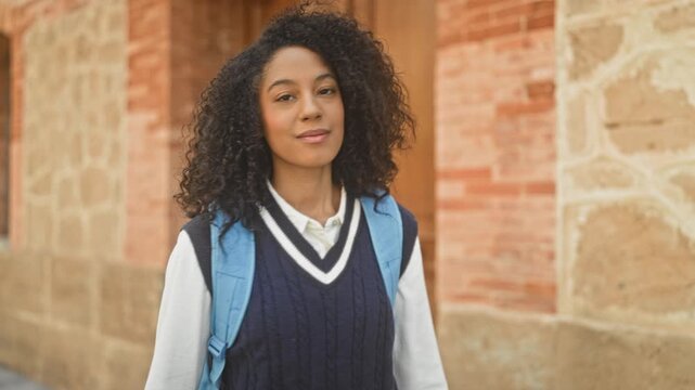 Young woman student with blue backpack crossing arms at brick building facade, steady gaze and slight smile; confidence.