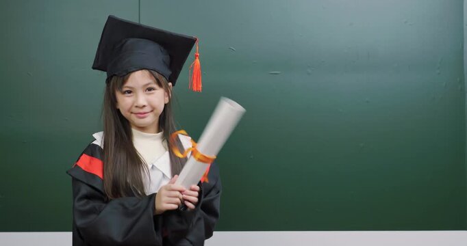 Happy Asian child in graduation gown holding scroll diploma, shaking it joyfully.