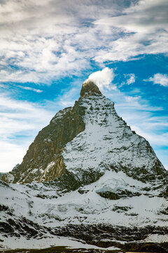 View of the iconic Matterhorn peak with its distinctive pyramid shape, snow-covered slopes, and a wispy cloud at the summit under a bright blue sky in Zermatt, Valais, Switzerland.